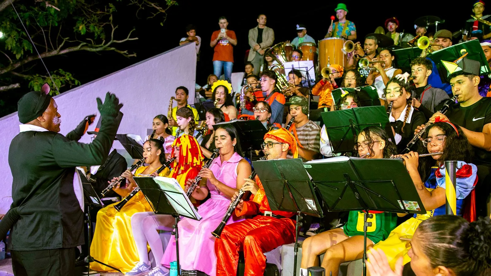 Banda de Música Mestre Pixuna em apresentação noturna: dezenas de músicos fantasiados tocam clarinetes, metais e percussão sob a regência do maestro.