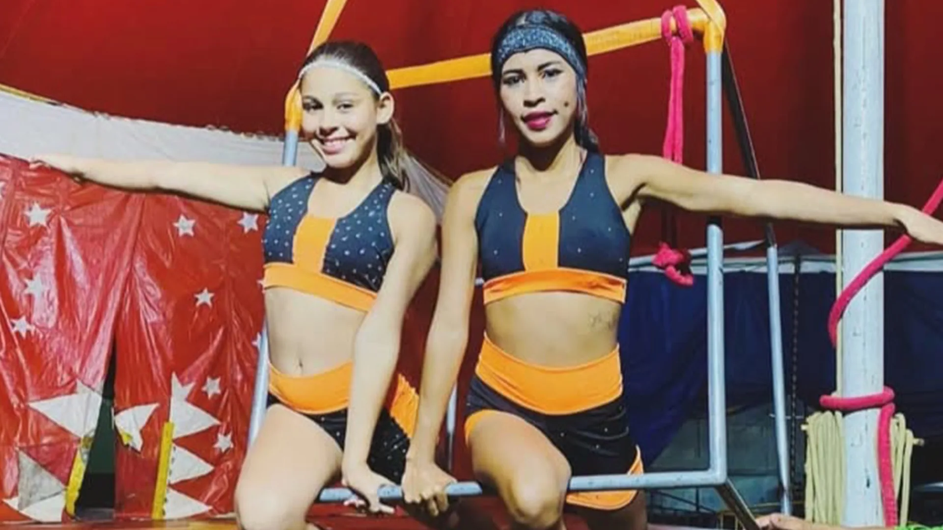 Bruna Bianca e Suyane Simões em cena: as duas artistas posam com os braços abertos sobre uma estrutura de barras, com lona de circo vermelha ao fundo
