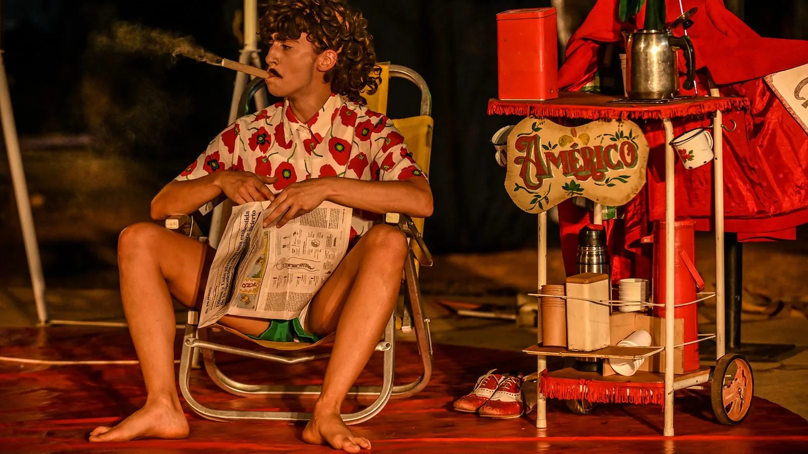Cami Basterra em cena de “Yo soy Américo”: palhaça de camisa florida sentada em cadeira de praia lendo jornal, ao lado de um carrinho vermelho de café com o letreiro “Américo”.