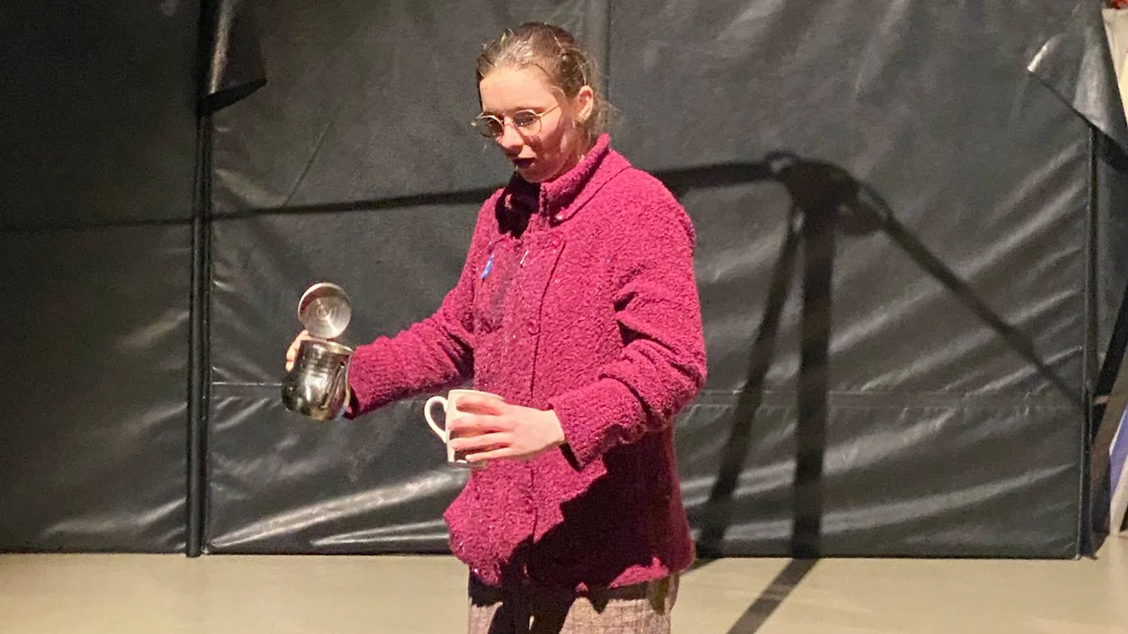 Camille Bekkers em cena de “Thébaïde”: performer de casaco magenta serve uma xícara diante de um fundo de lona preta.