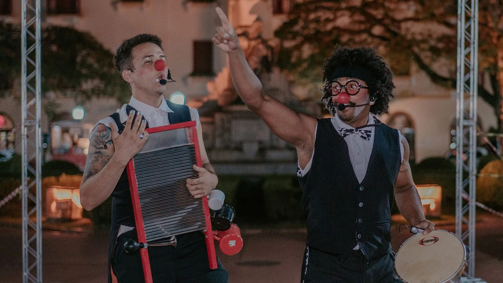 Companhia Penduricalhos em cena: dois palhaços músicos — um com tábua de lavar roupa, outro com tamborim — em apresentação noturna.