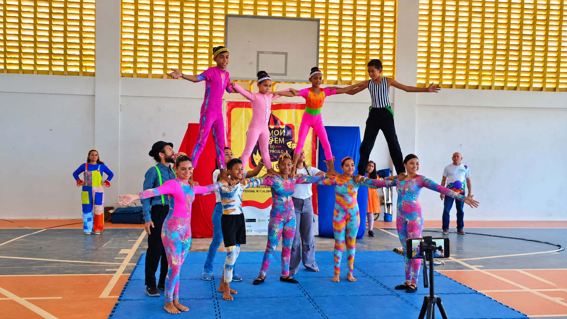 Circo Escola Canoa Criança em cena: crianças e adolescentes com macacões coloridos formam uma pirâmide humana sobre tatames azuis em uma quadra de esportes, com quatro jovens equilibrados nos ombros dos colegas e braços abertos, sob a parede de tijolos amarelos vazados.