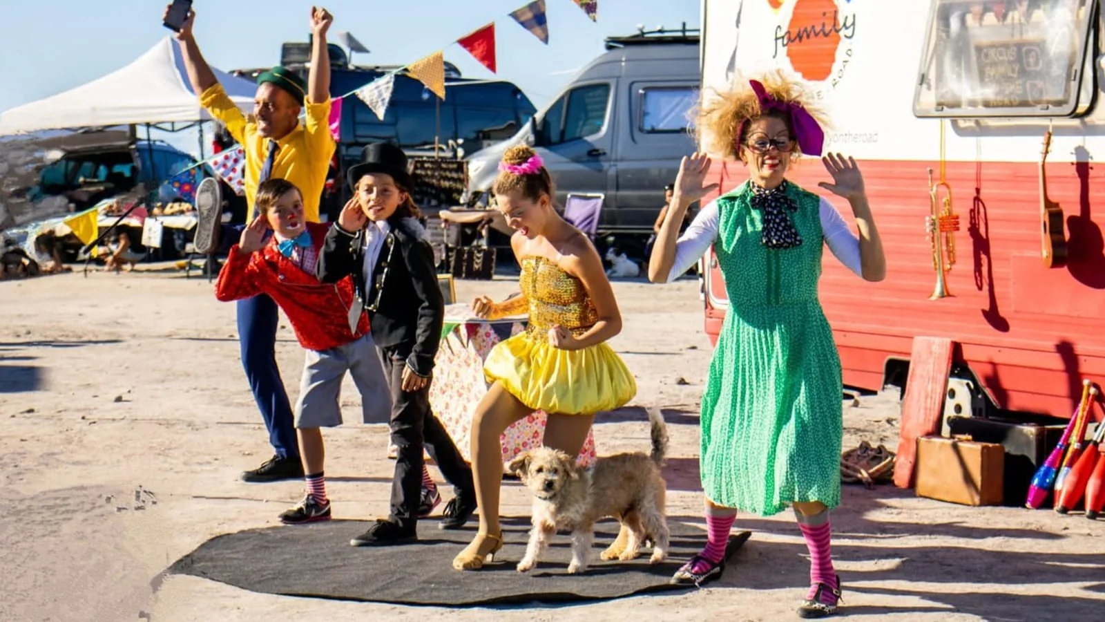 Circus Family On The Road em cena: família de artistas circenses e um cachorro diante de um trailer vermelho, em terreno ensolarado.