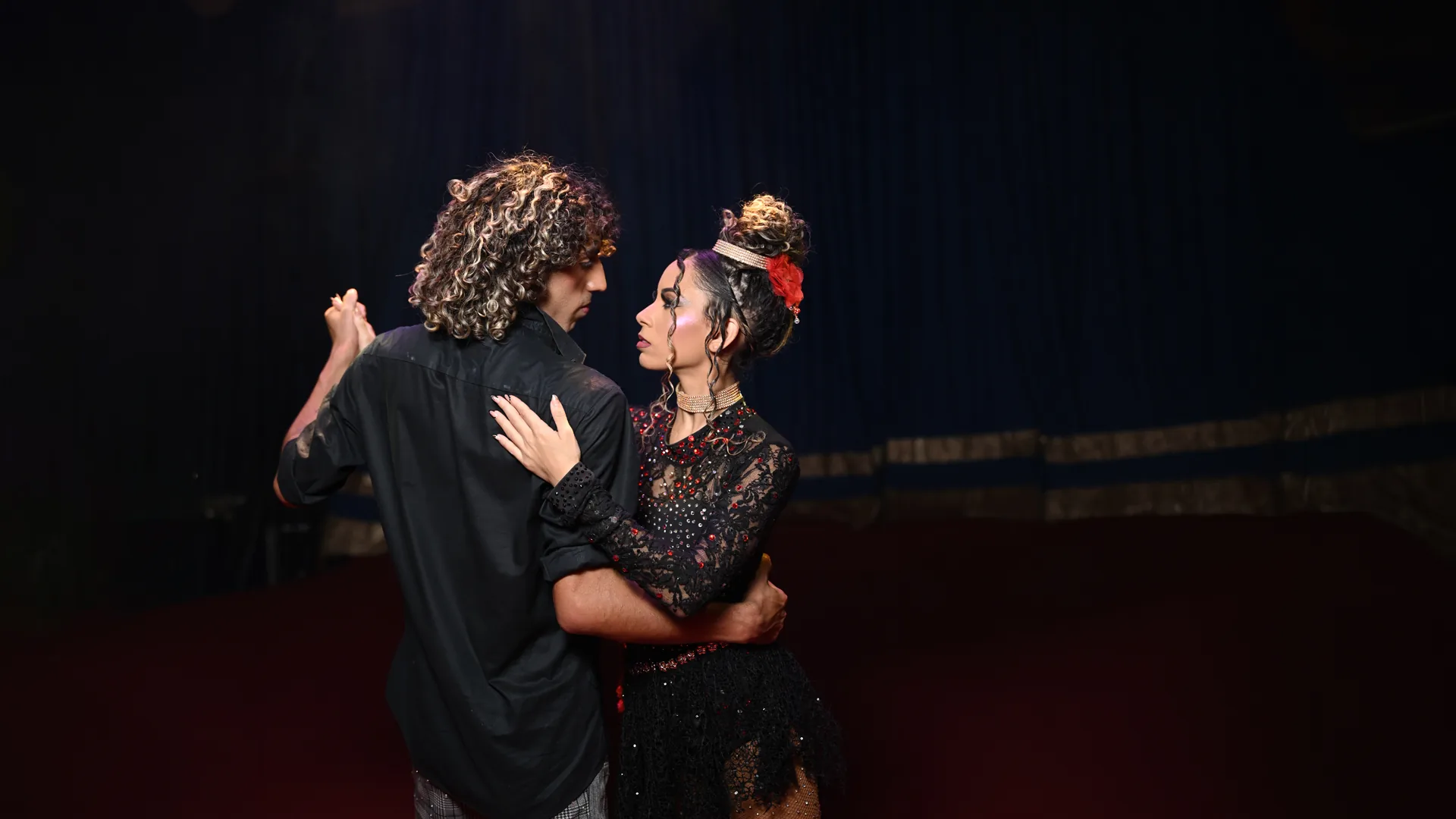 Duo Aéreo Ícaro em cena de “A bailarina que sonha em se casar”: o casal dança abraçado no picadeiro — ele, de costas, com cabelos cacheados e camisa preta; ela, de vestido de renda preta com brilhantes, gargantilha de pérolas e flor vermelha no coque —, sob fundo de cortina escura.