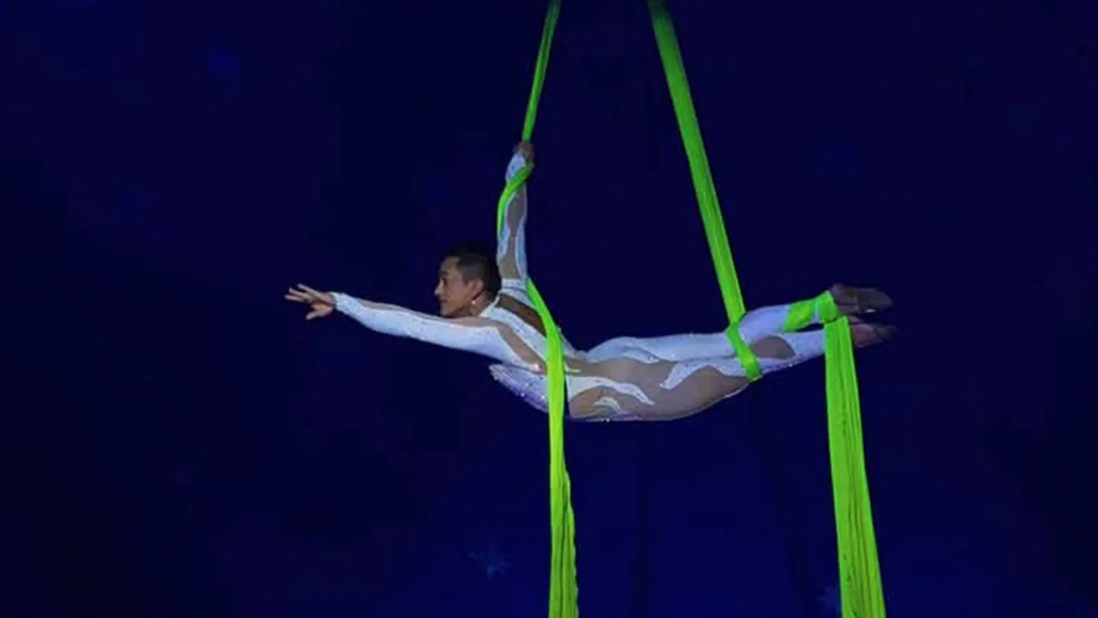 Erick Ronaldy em número aéreo: o artista estende o corpo na horizontal, suspenso por tecidos verde-limão, contra o fundo azul-escuro.