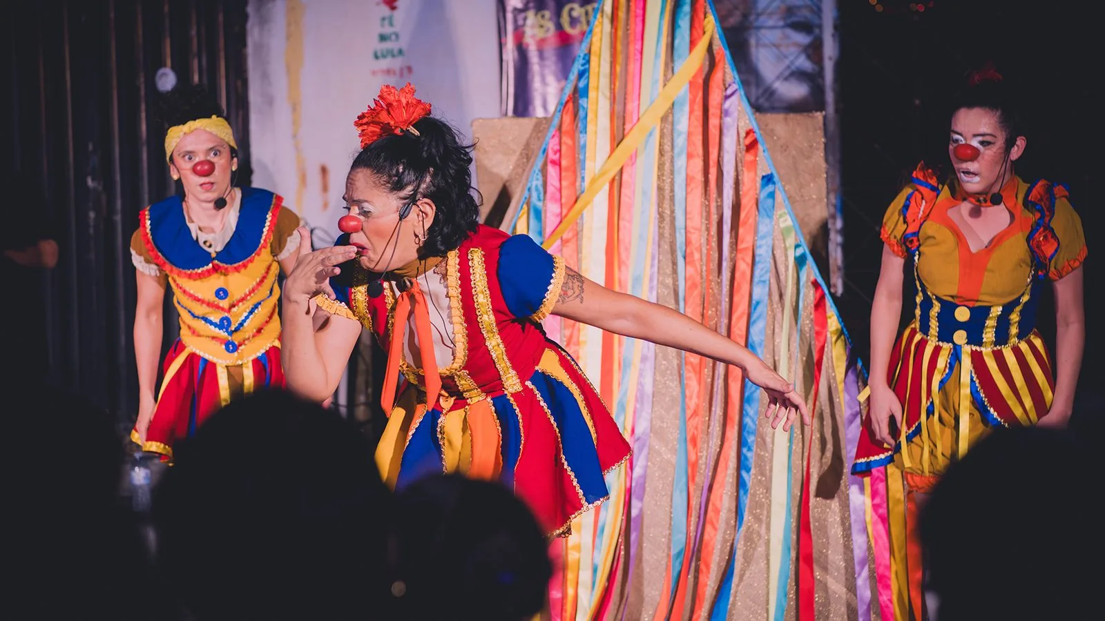 Grupo Garajal em cena de “As Circas”: três palhaças em figurinos de listras coloridas e babados, junto a fitas multicoloridas.