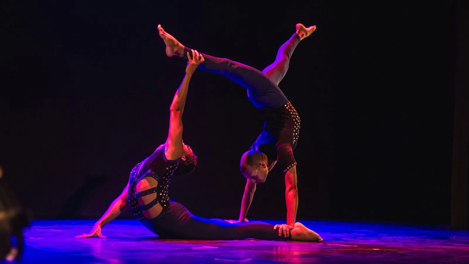 Ítalo Feitosa em cena: dupla de acrobacia — um artista em parada de mão é sustentado pelo parceiro em posição de arco, sob luz magenta e azul.