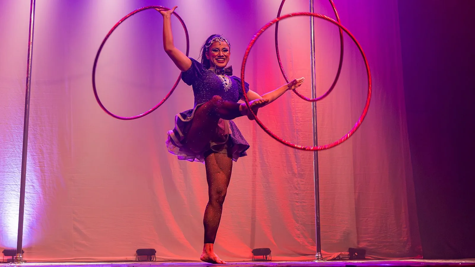 Laísa Fonseca em cena: a artista equilibra-se em uma perna só girando três bambolês, sob luz magenta de palco.
