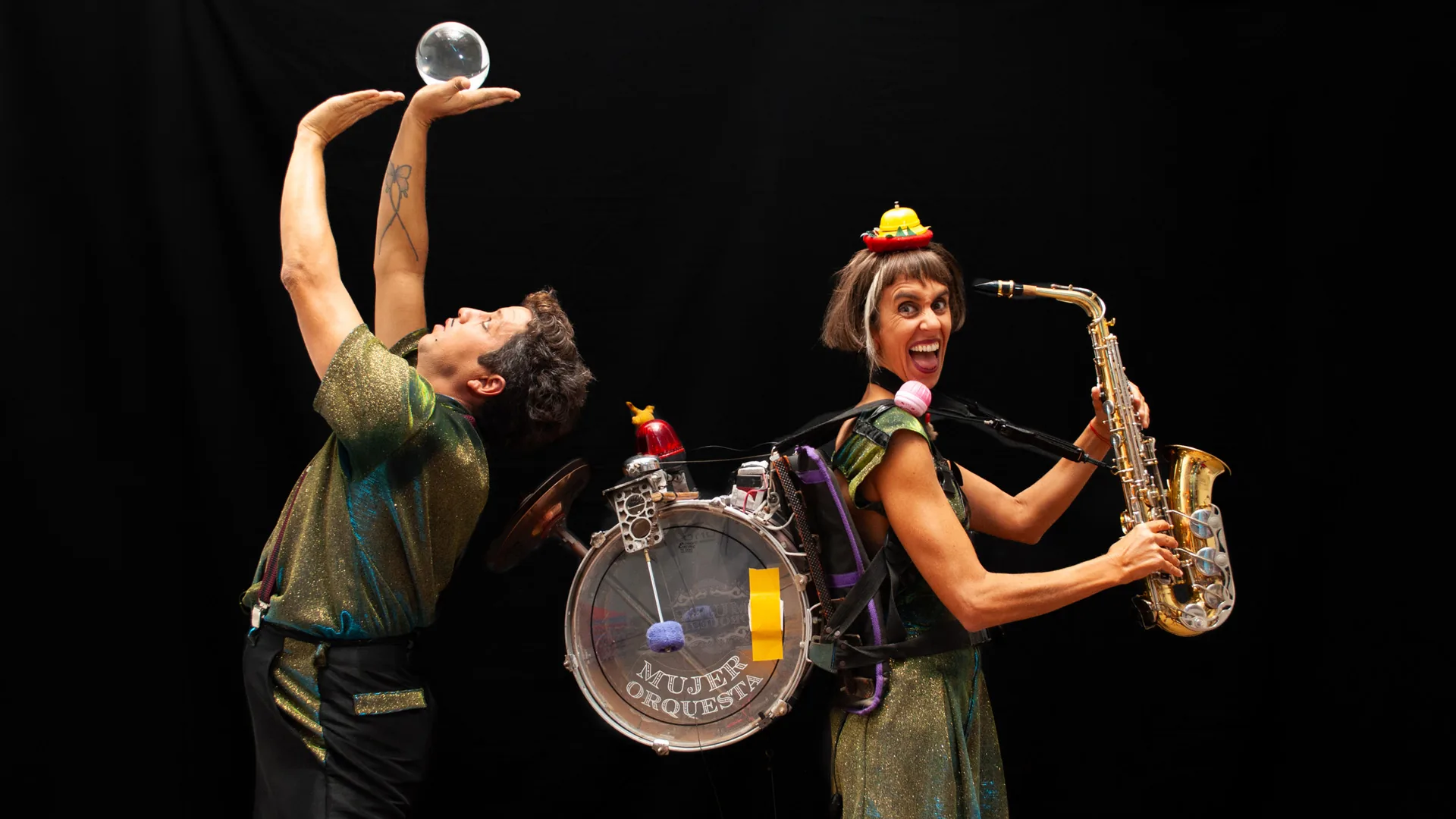 Latin Duo em cena de “Se desconcierta el concierto”: o artista equilibra uma bola de cristal sobre a mão erguida enquanto a parceira, de chapéu-bule miniatura e bateria presa ao corpo com a inscrição “Mujer Orquesta”, toca saxofone, contra o fundo negro.