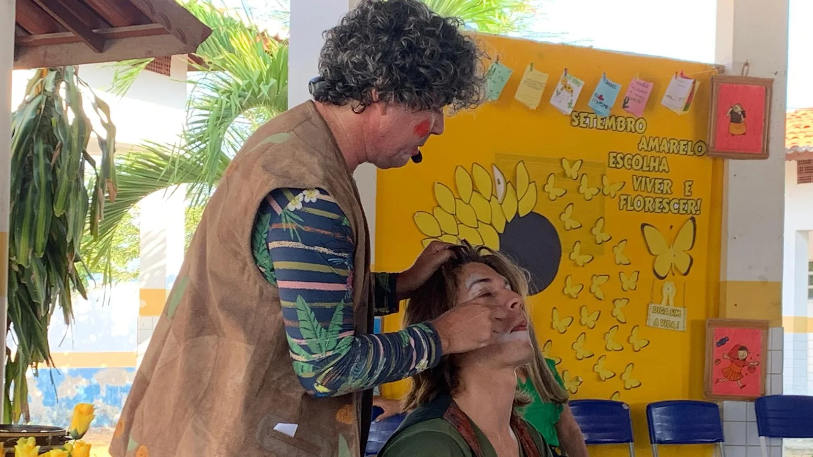 Orlângelo e Artur em ação do FICC nas Escolas: palhaço maquia uma participante diante de um mural escolar amarelo.