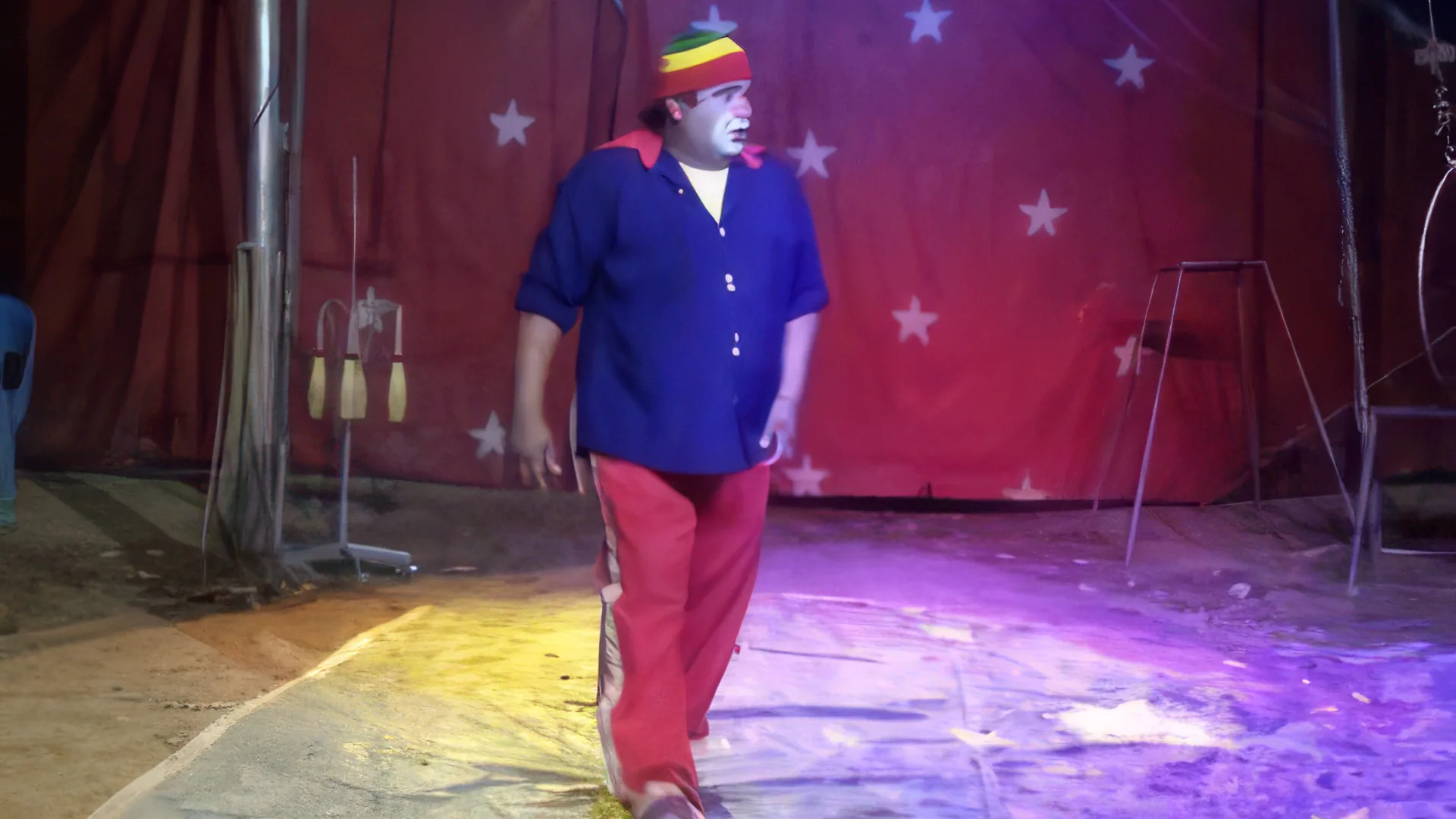 Palhaço Soneca em cena no picadeiro: de rosto branco, gorro listrado em verde, amarelo e vermelho, camisa azul e calça vermelha, ele caminha sobre o chão iluminado em amarelo e roxo, diante da lona vermelha estampada com estrelas brancas.