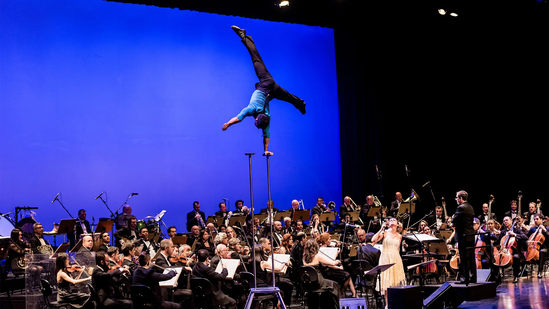 Paulo Maêda em cena de “Em prumo”: o artista faz uma parada de mãos com uma só mão sobre hastes altas, com as pernas abertas em tesoura, acima de uma orquestra completa em palco com fundo azul, regente e uma cantora de vestido claro.
