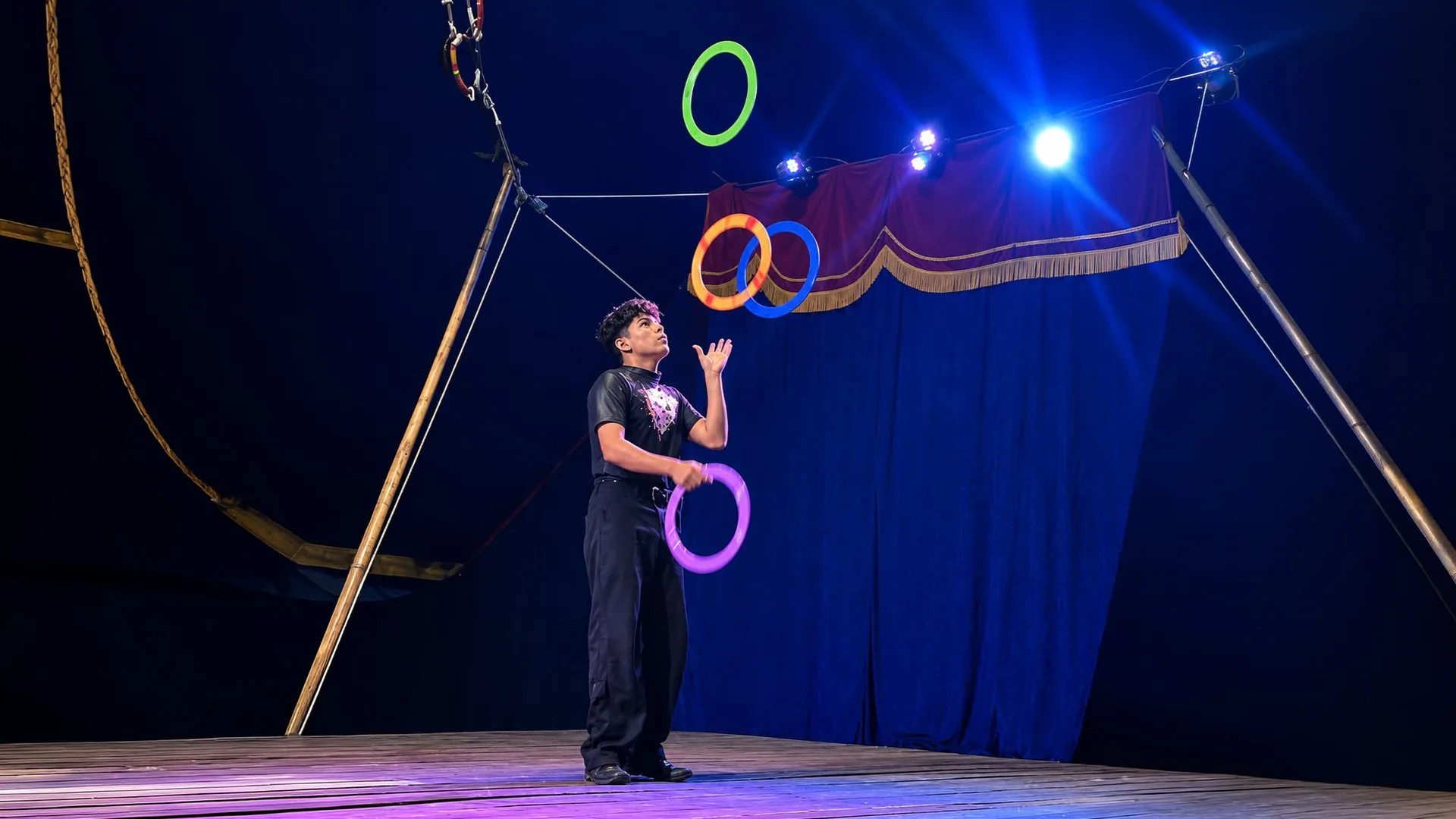 Reynaldo em cena no Show de Malabares: o artista lança ao alto aros coloridos — verde, laranja, azul e roxo — diante da cortina azul do picadeiro