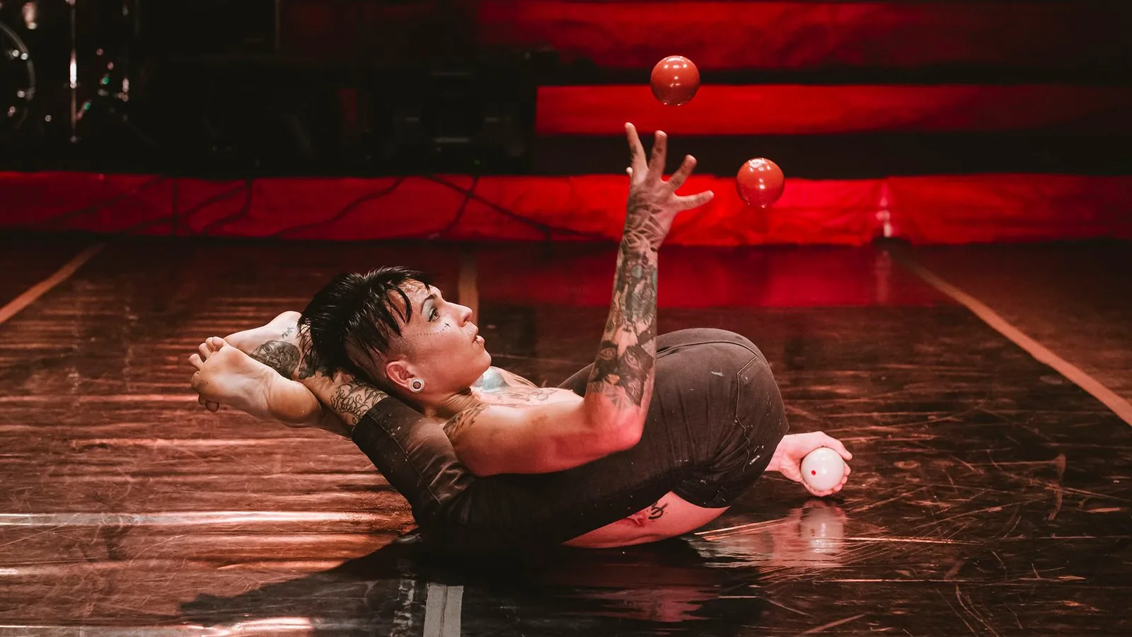 Yako em cena de “Experimento Yako”: artista em posição de contorsão no chão do picadeiro faz malabarismo com bolas vermelhas.